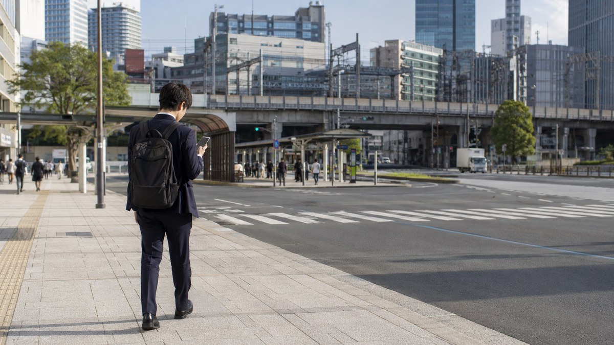 駅前でスマートフォンを見ながら歩く若いビジネスマン。都市部では車を持たない若者が増えている