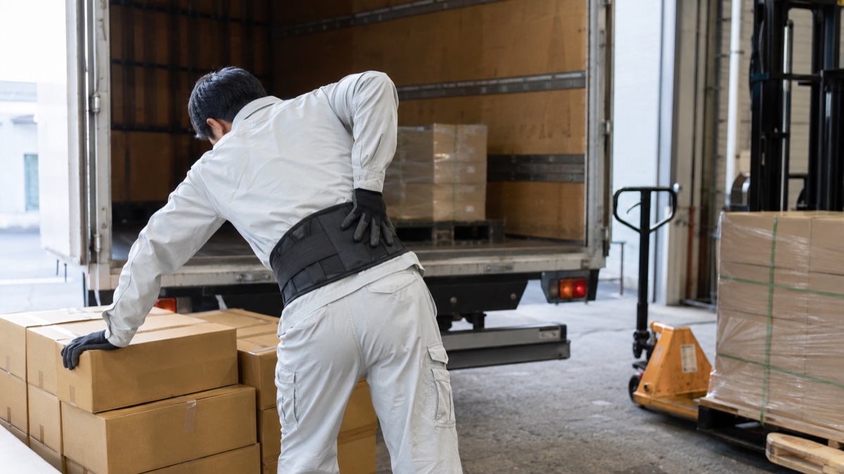 腰痛ベルトを巻き、荷物の積み下ろしで腰に負担を感じているトラックドライバー